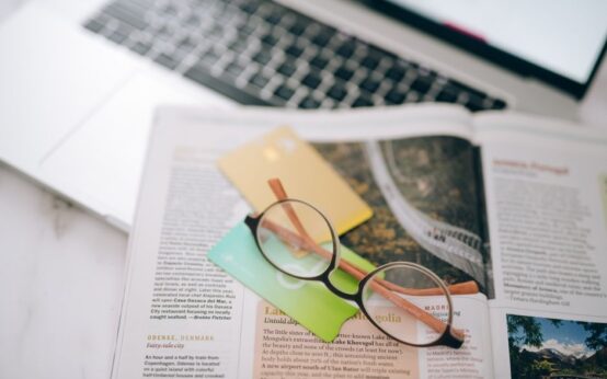 Overhead view of reading glasses, credit cards, and a magazine with a blurred laptop in the background.