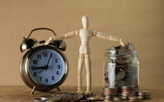 Wooden mannequin holding an alarm clock and a jar full of coins, symbolizing time and money.